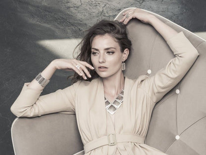 Mulher elegante reclinada numa poltrona usando pulseira hedra prata feminina, colar geométrico, brincos e anéis